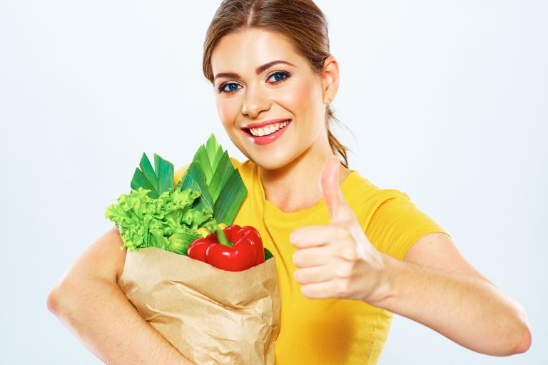 A lady holding a paper bag overflowing with fresh vegetables. She's giving a thumbs up.