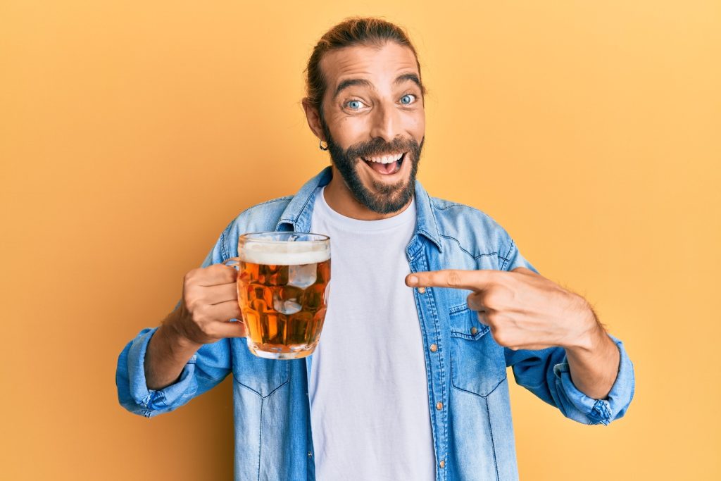 A happy, smiling man holding a glass full of beer and pointing to it.