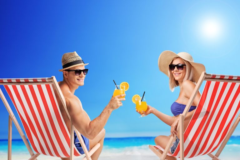A couple sitting on a beach in striped chairs, Holding cocktails. They are both wearing hats and sunglasses.