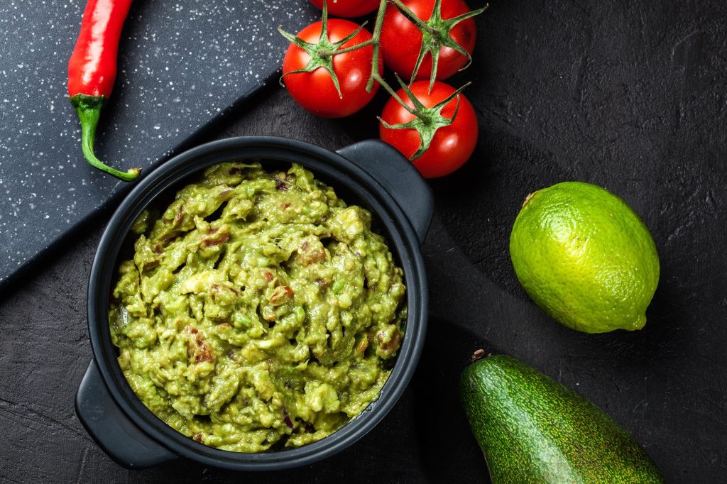 A black bowl of guacamole.  Chipotle, tomatoes, lime and jalapenos beside the bowl.