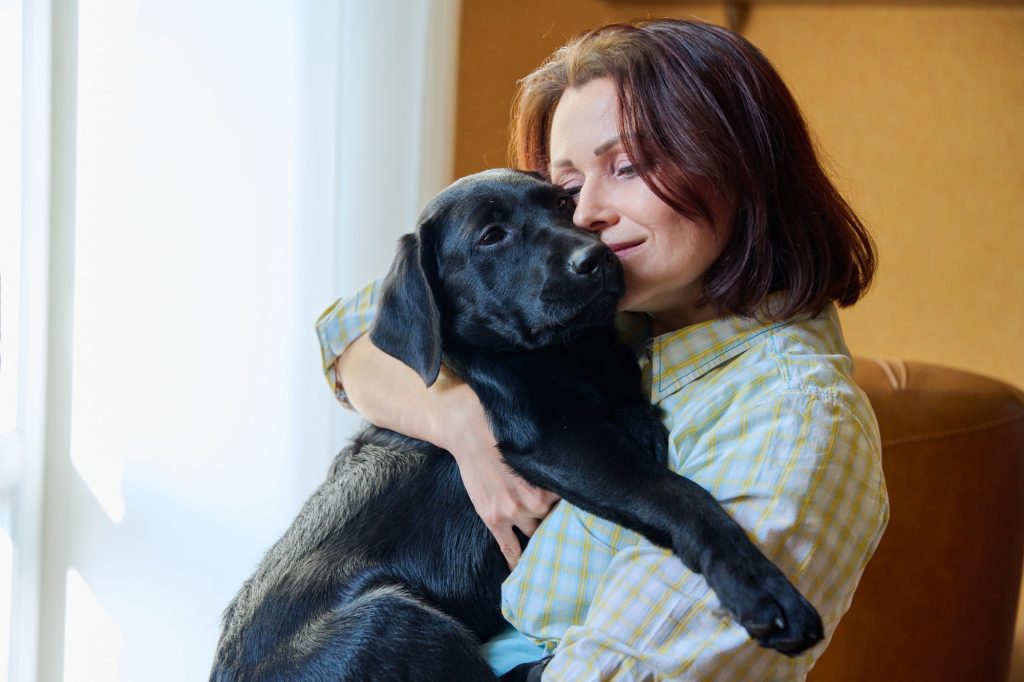 A middle age brunette woman hugging a black middle size dog.