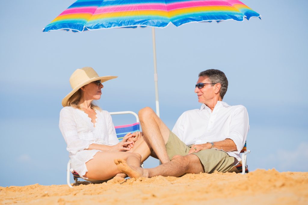 A man and woman sitting on a beach under a striped colorful umbrella.