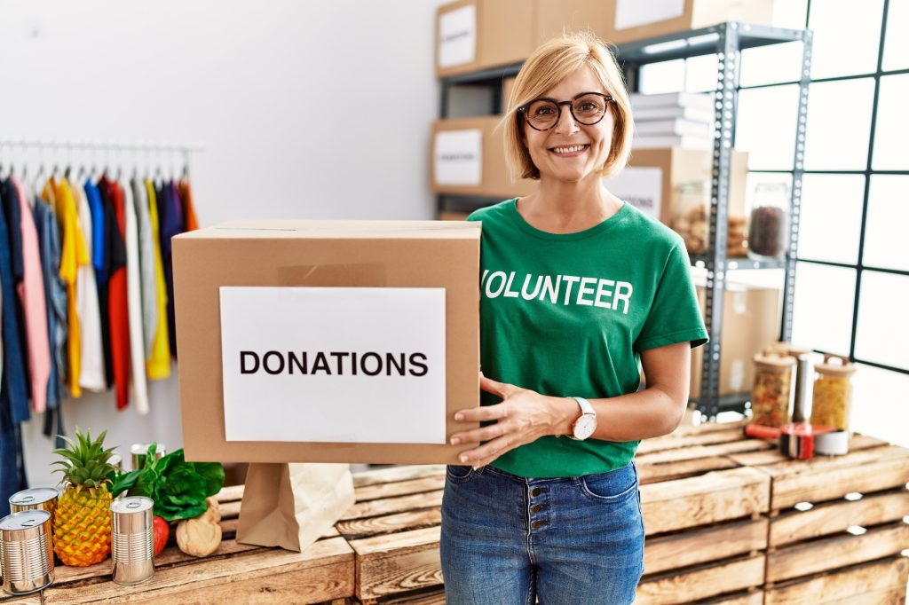 A lady with a shirt on that reads, volunteer.  She's holding a box that reads, donations.