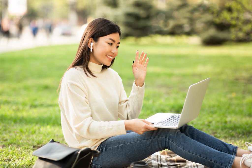 A college student on a video chat on laptop, sitting outside on the grass.