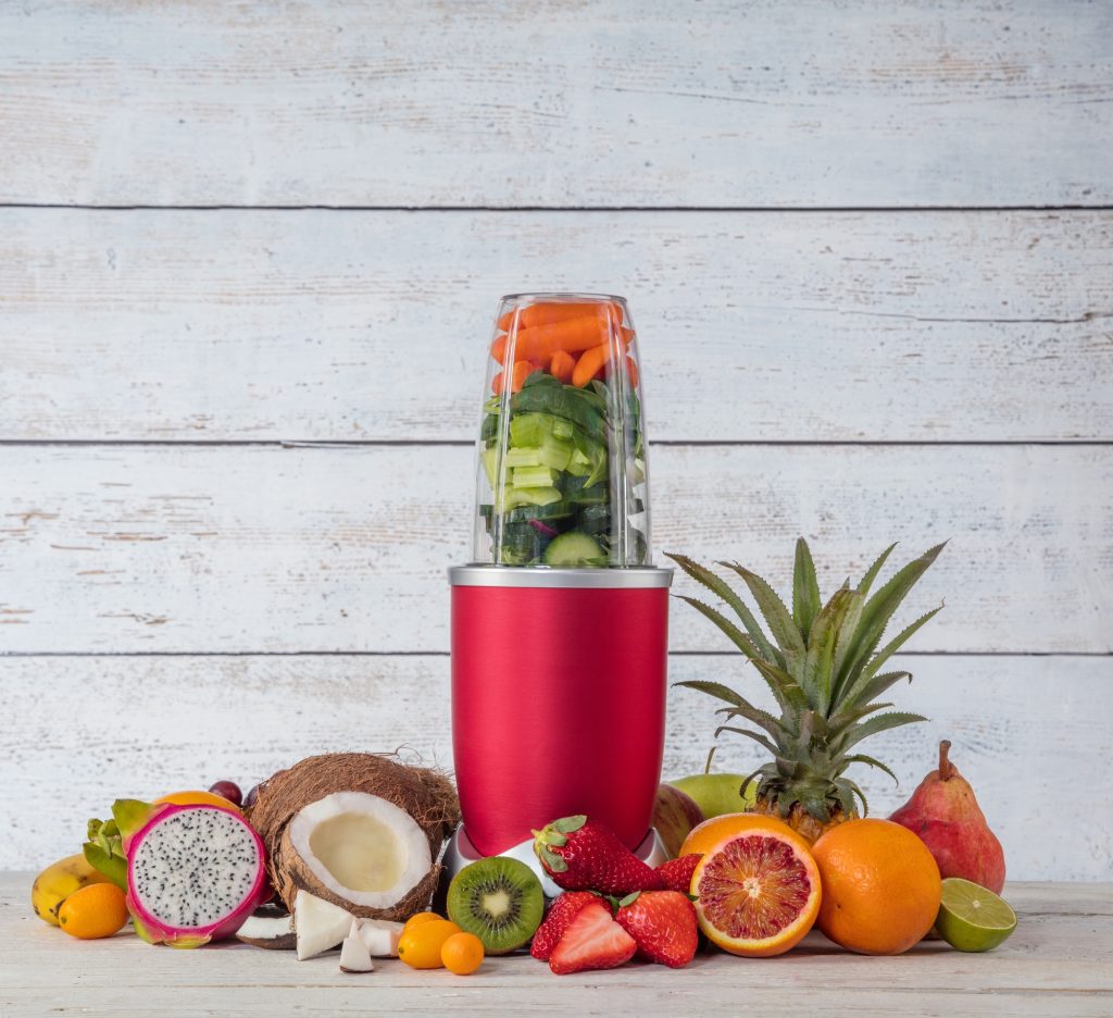 A red personal blender filled with and surrounded by a variety of fruits.