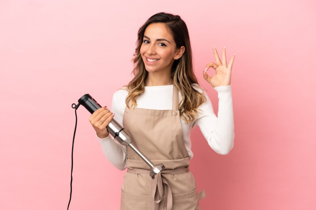 Girl holding a immersion blender giving the a okay signal with other hand.