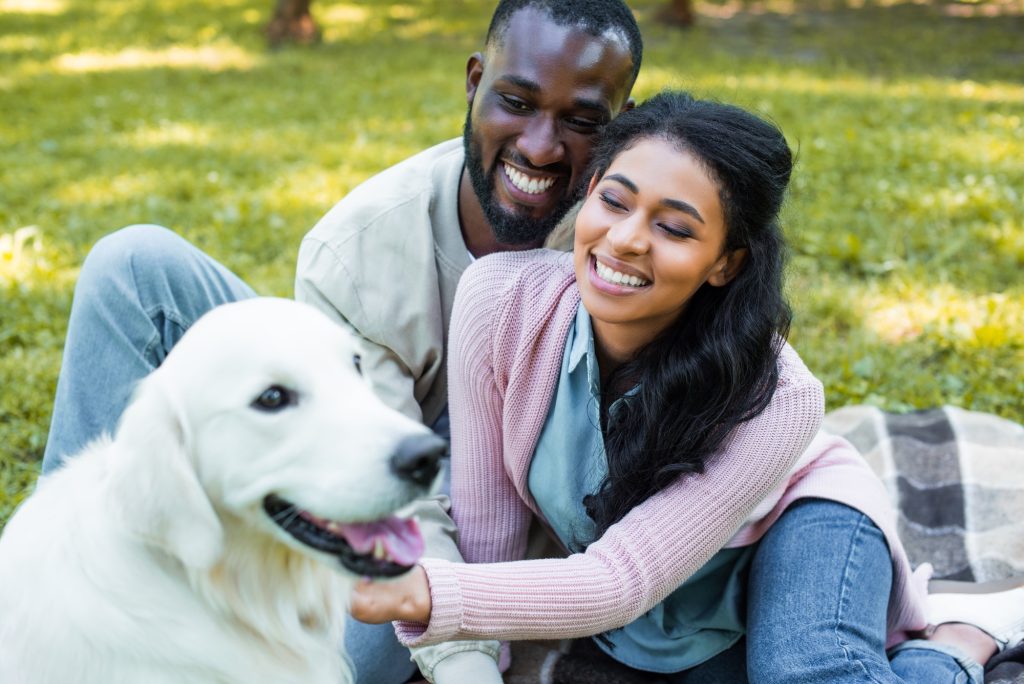 A man and woman smiling, sitting outside petting a large white dog.