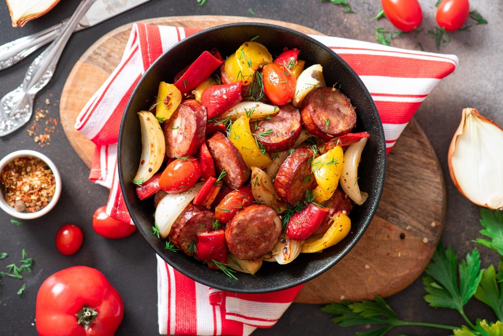 Sausage and peppers cooked in a black skillet.  Laying on a red and white cloth.  Ingredients surrounding the skillet.