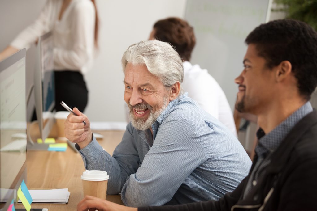 Older man at work smiling, looking at a computer screen with coworkers.