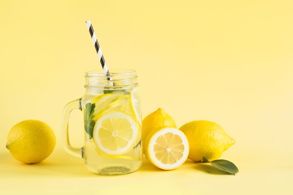 A Clear glass with lemonade and lemons in it and a striped straw sticking out of it.  The background is yellow. There are lemons around it.