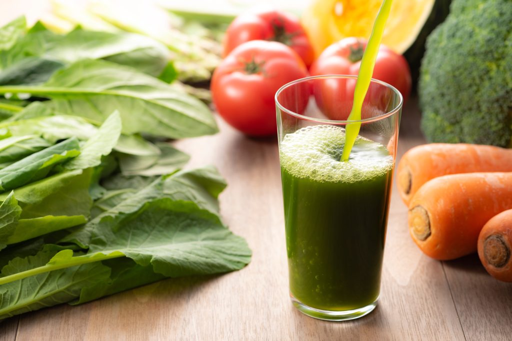 A glass of green vegetable juice and vegetables all around it.