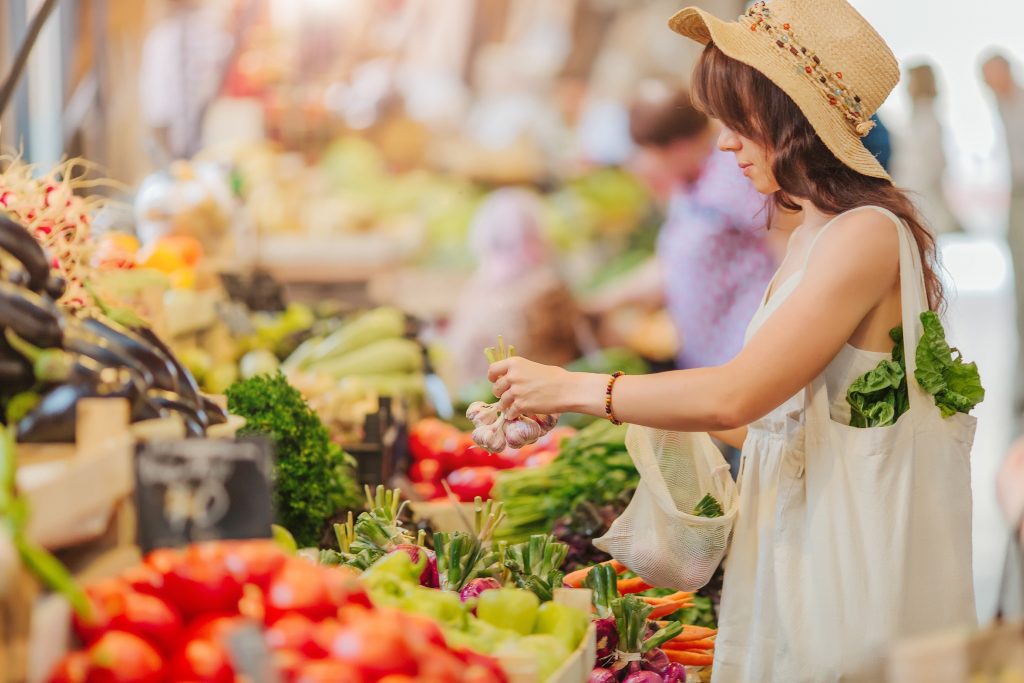 A woman with a hat and shopping bag shopping at a farmers market.