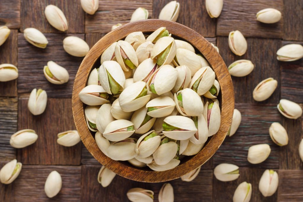 A wood bowl filled with pistachios, and they are scattered around it.