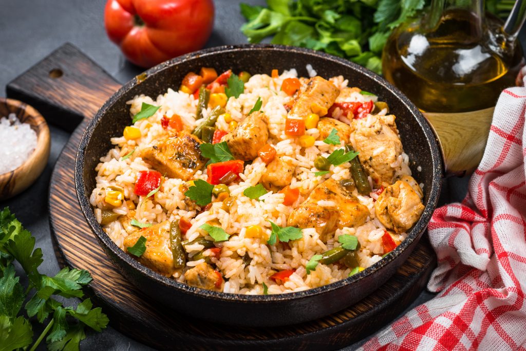 A black bowl filled with chicken rice and veggies. The bowl is surrounded by fresh vegetables.