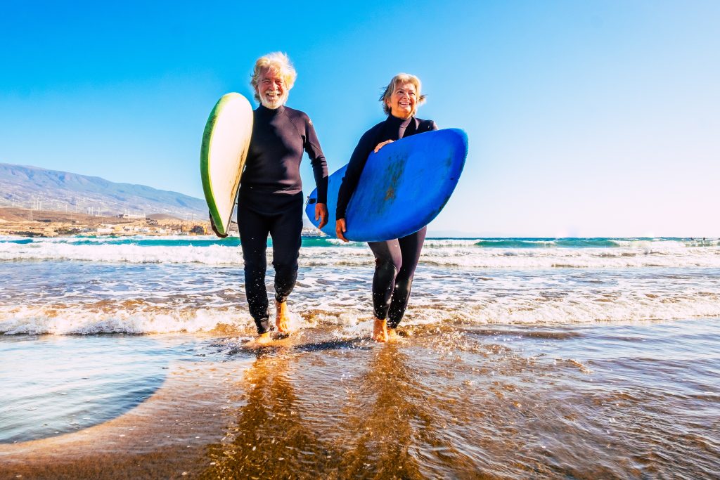 Older man and woman, both holding surf boards walking out of the water.