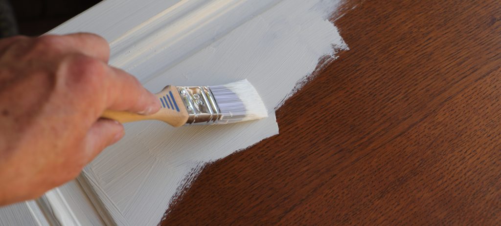 a hand holing a paint brush, brushing white paint on a dark wood cabinet door.