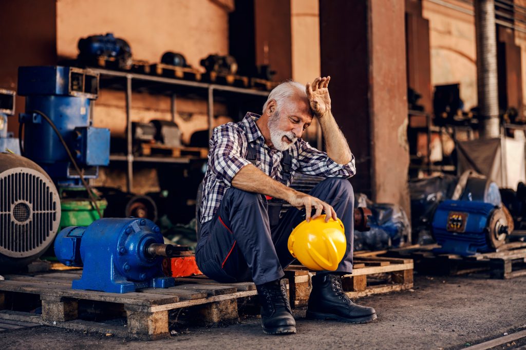 An exhausted old man sitting in a factory with his head in his hand.  He's holding a hard hat.