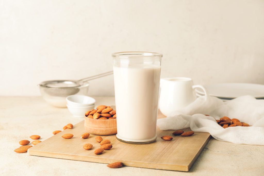 A clear glass with almond milk in it sitting on a wooden board. Almonds are scattered around the glass.