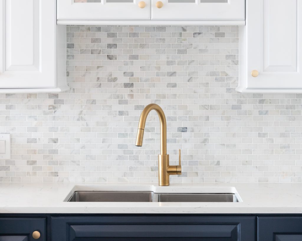 Kitchen sink, gold faucet and gray and white tile backsplash.