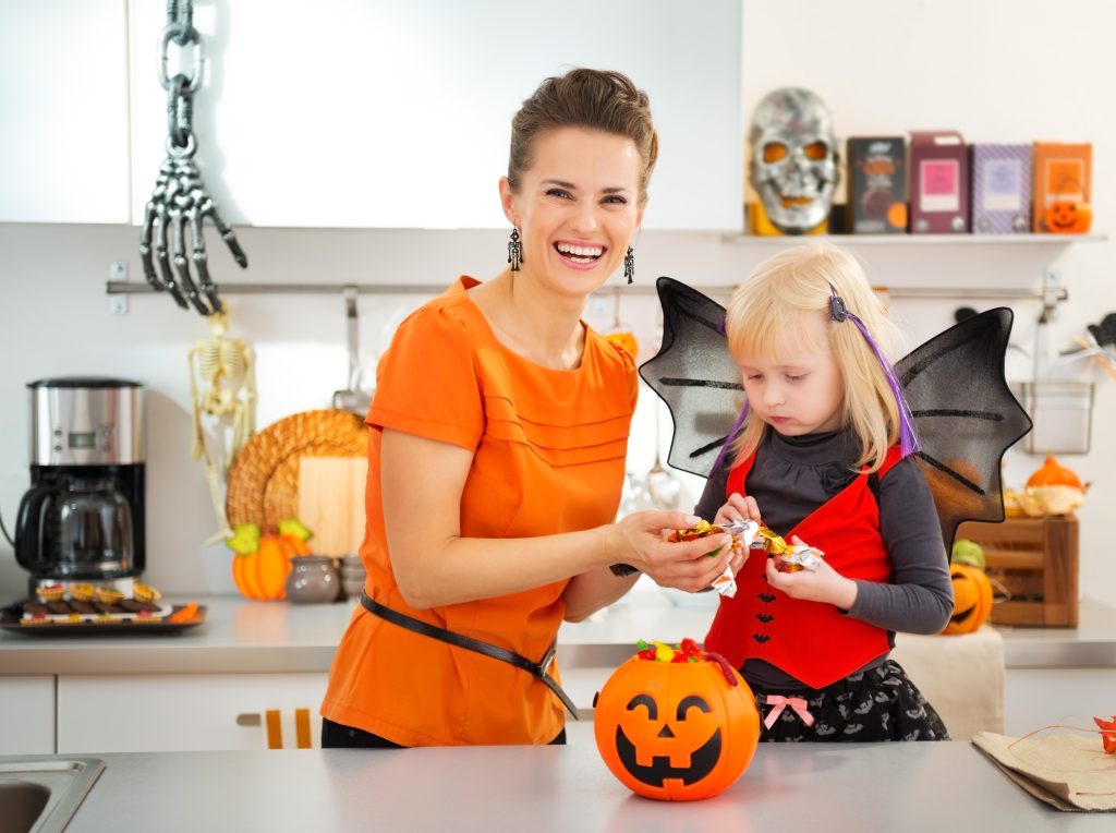 A mom and child holding a pumpkin container filled with Halloween candy. They are in a kitchen.