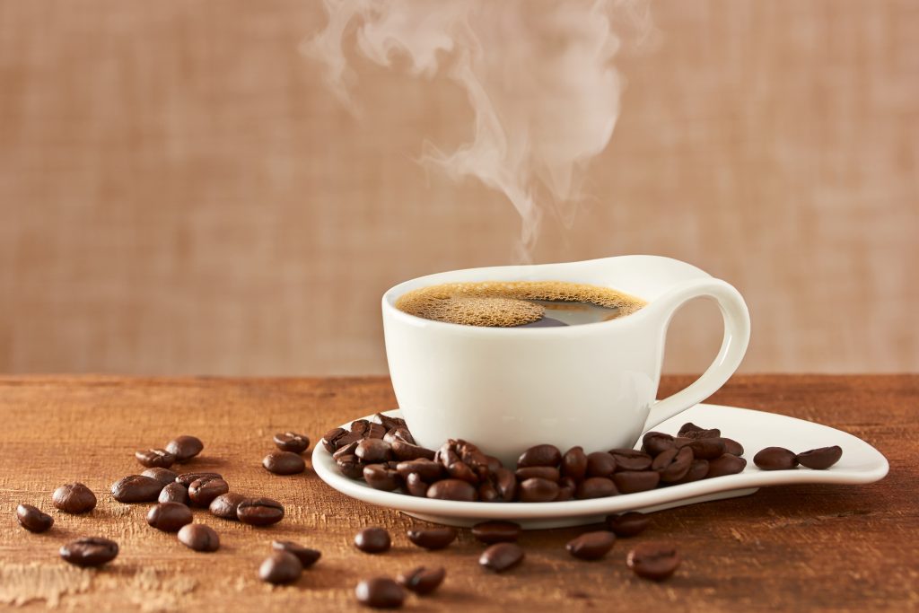 A white cup of hot coffee with steam rising from it.  coffee beans all around the cup.