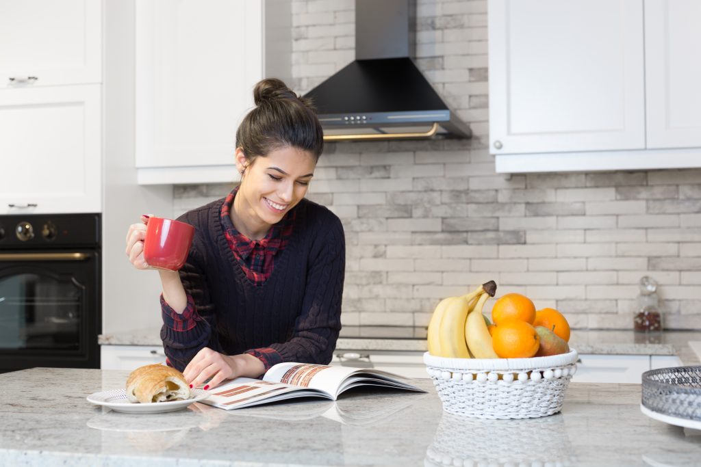 A woman in kitchen holding a mug and reading a magazine.