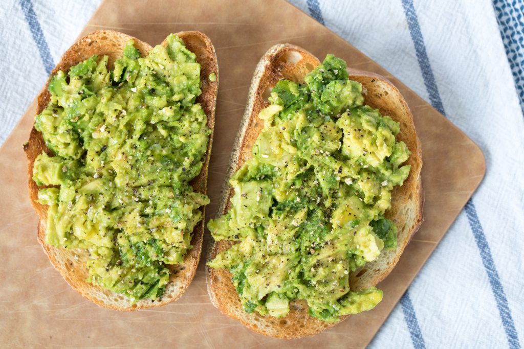 Two slices of toast with mashed avocado on brown parchment paper.