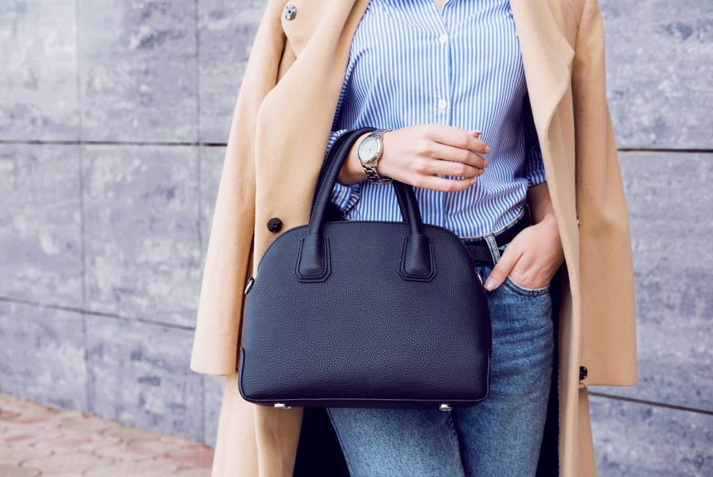 pictured bottom half of a womans fashion photo, wearing a tan wool coat, jeans, pinstripe shirt and holding a leather handbag.
