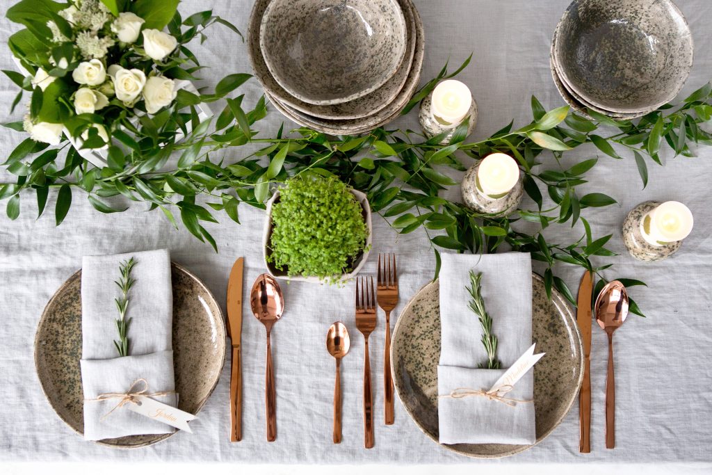 Overhead view of an elegant table setting, neutral colors and greenery, the flatware is bronze, the dinnerware is beige.