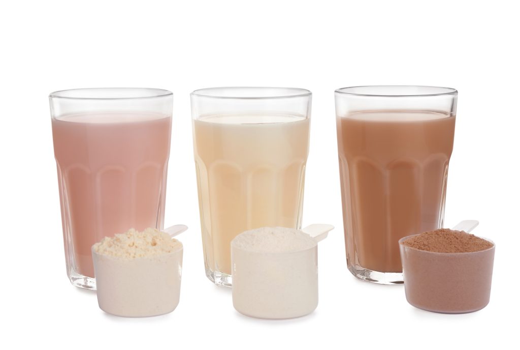 3 glasses with different flavors protein shakes and 3 scoops of protein powder beside them.