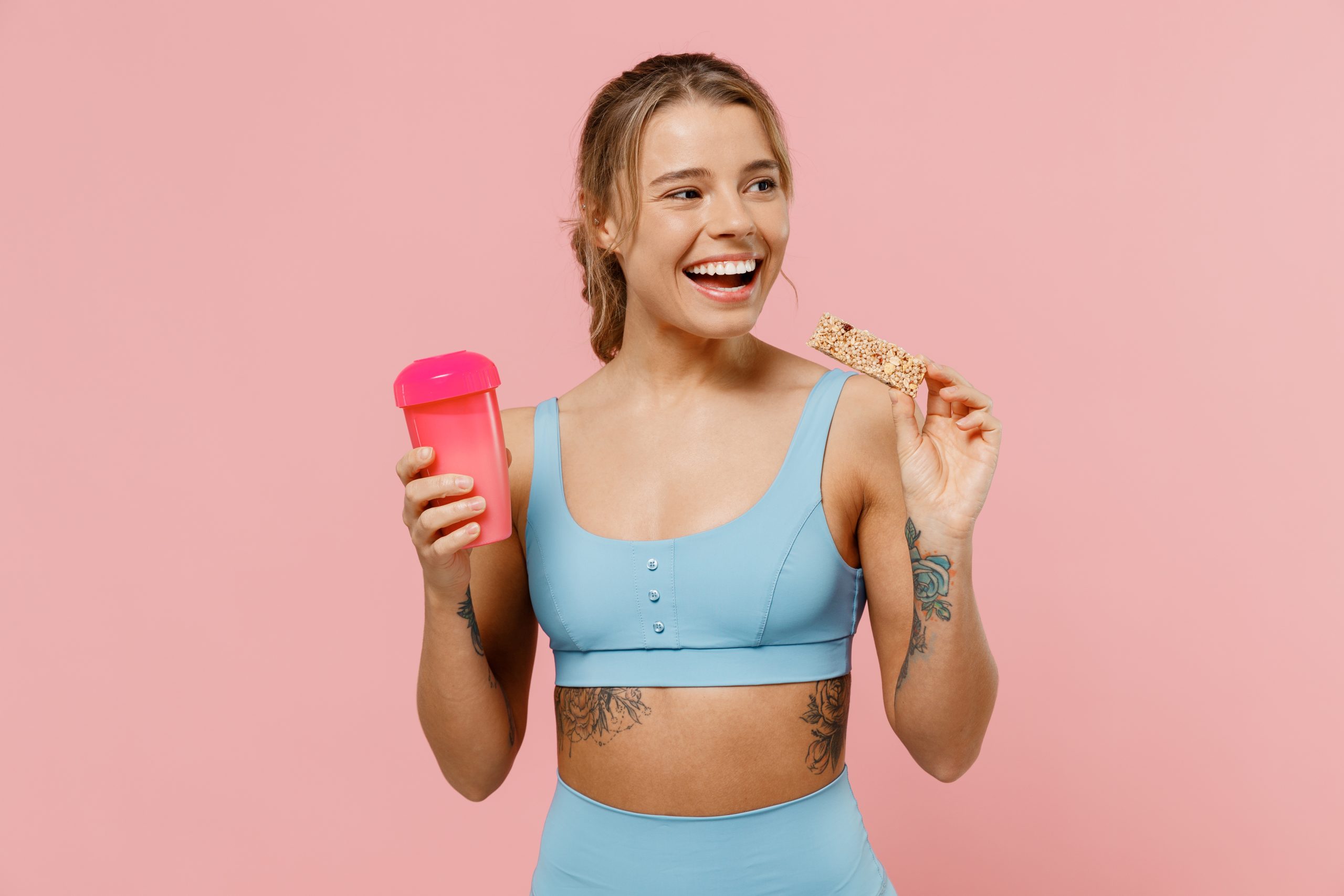 A woman wearing fitness wear, holding a protein shake and a protein bar.