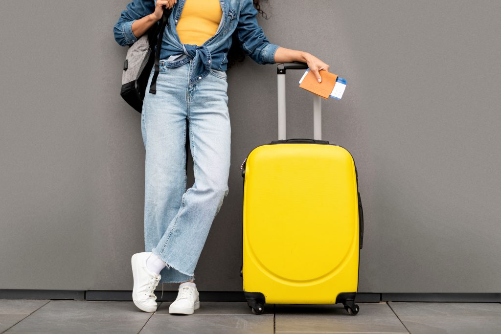 unrecognizable person standing beside a yellow suitcase, holding a passport.
