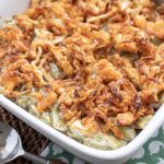 green bean casserole in a white dish. a wood handled spoon beside it. sitting on a green and white napkin.