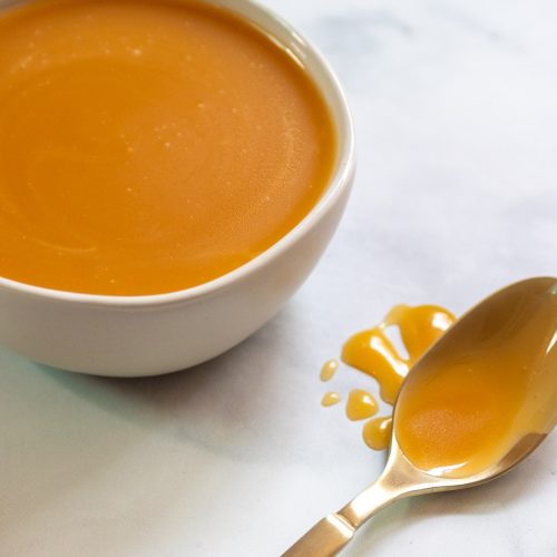 caramel sauce in a white bowl with a gold spoon covered in caramel sauce.