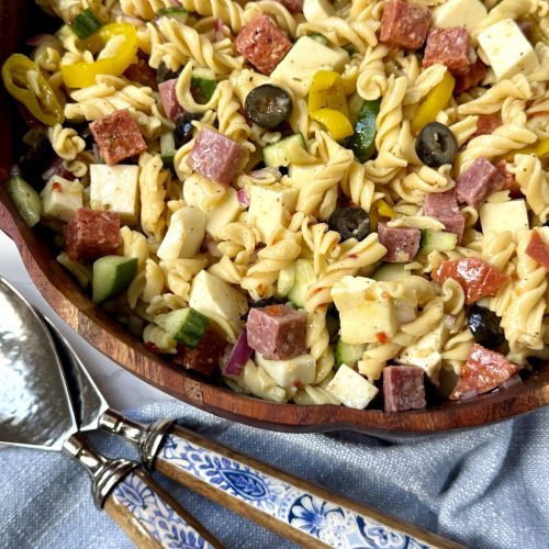 pasta salad in a wooden bowl. blue and white floral salad tongs, a blue cloth napkin.
