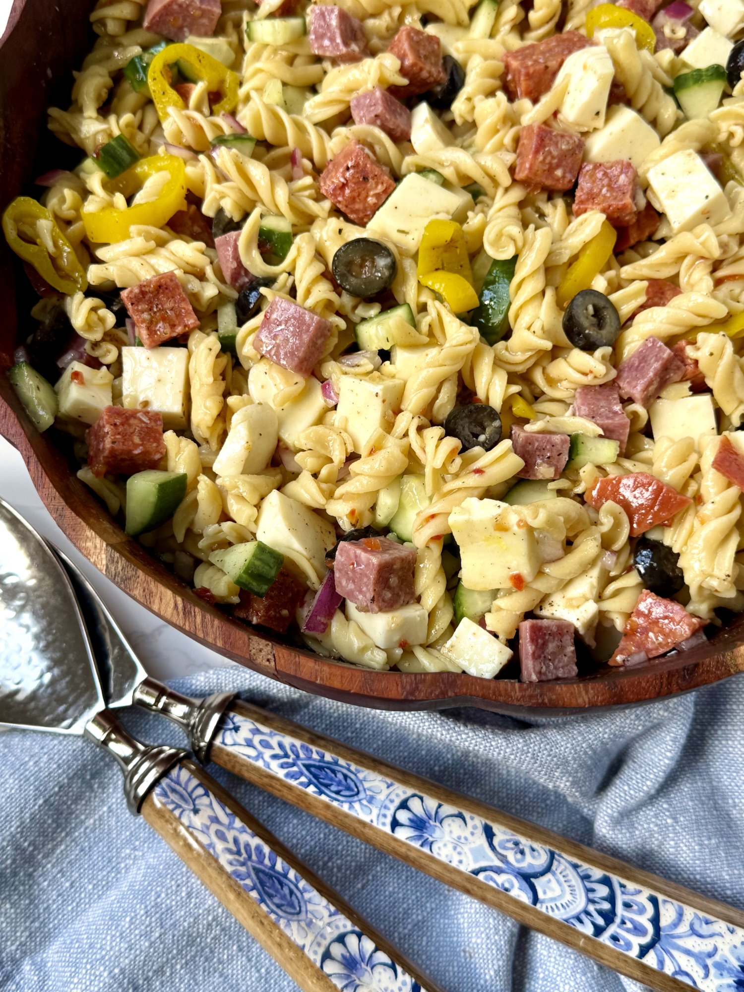 pasta salad in a wooden bowl. blue and white floral salad tongs, a blue cloth napkin.