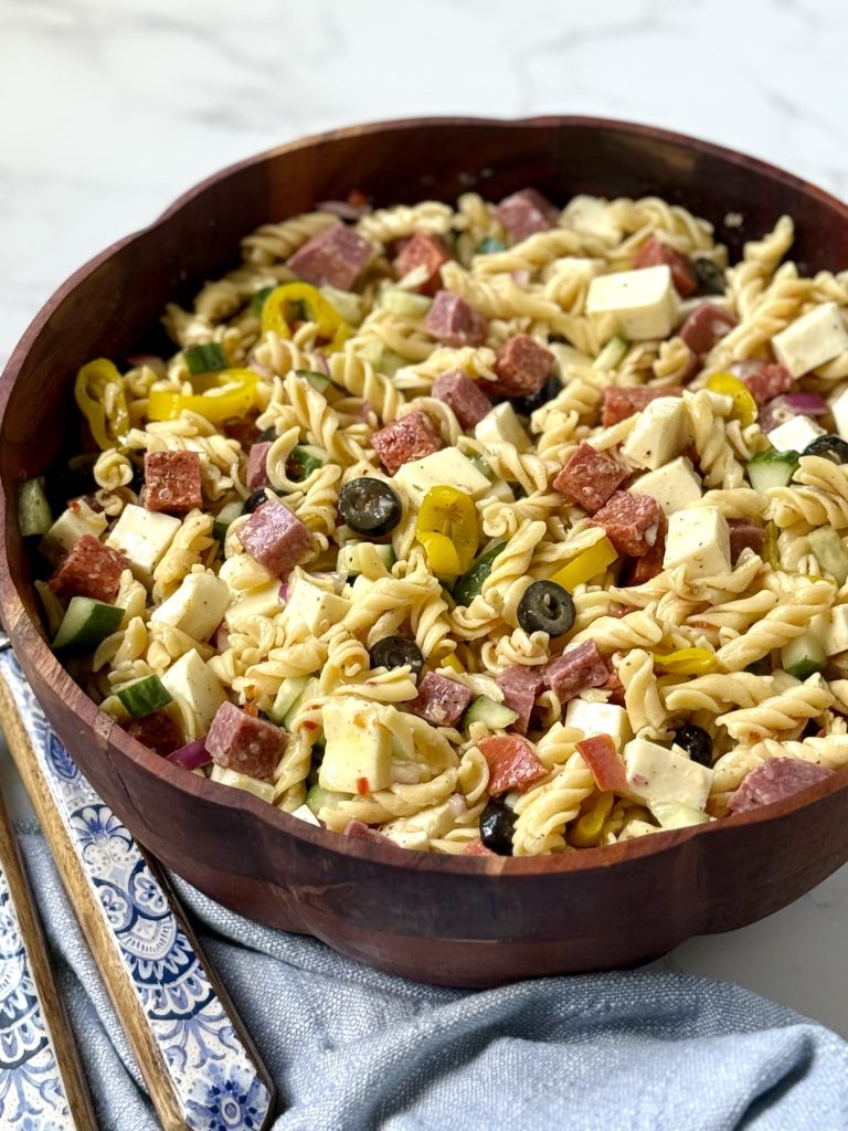 pasta salad in a wooden bowl. blue and white floral salad tongs, a blue cloth napkin.