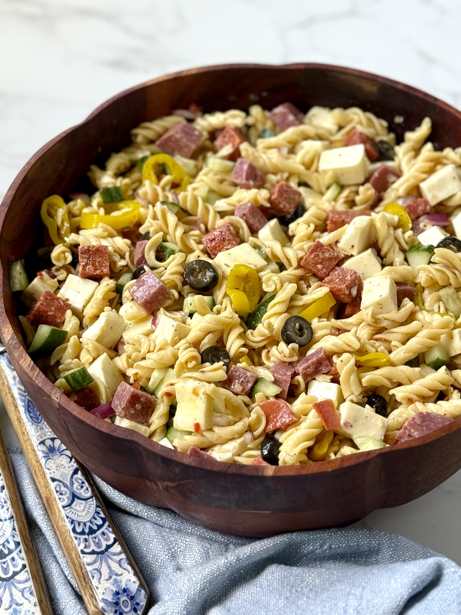 pasta salad in a wooden bowl. blue and white floral salad tongs, a blue cloth napkin.